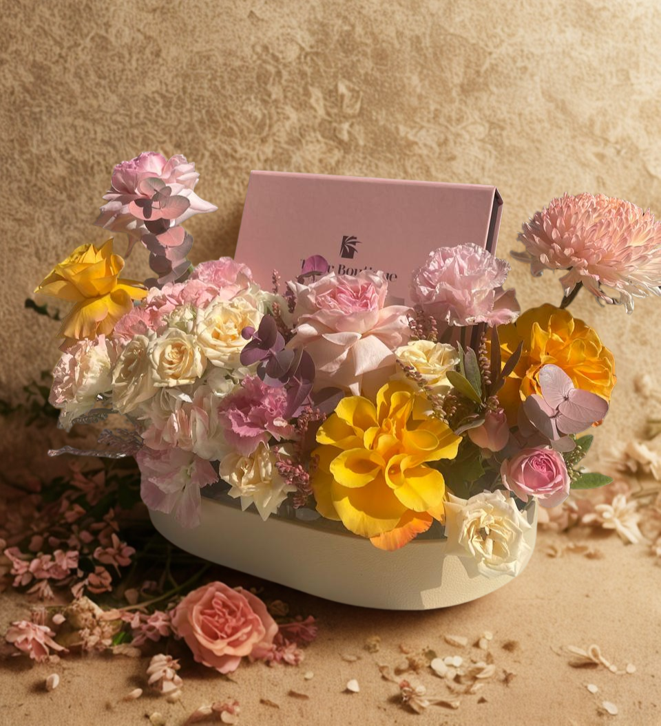 Floral arrangement in a heart-shaped box with a pink card on a beige background with a box of chocolate dates to gift